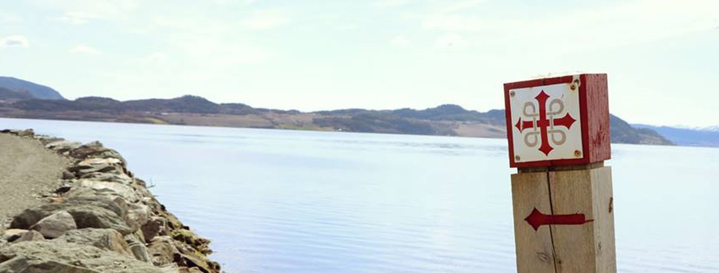 Bilden visar en stolpe med S:t Olavsledens logga med vatten i bakgrunden